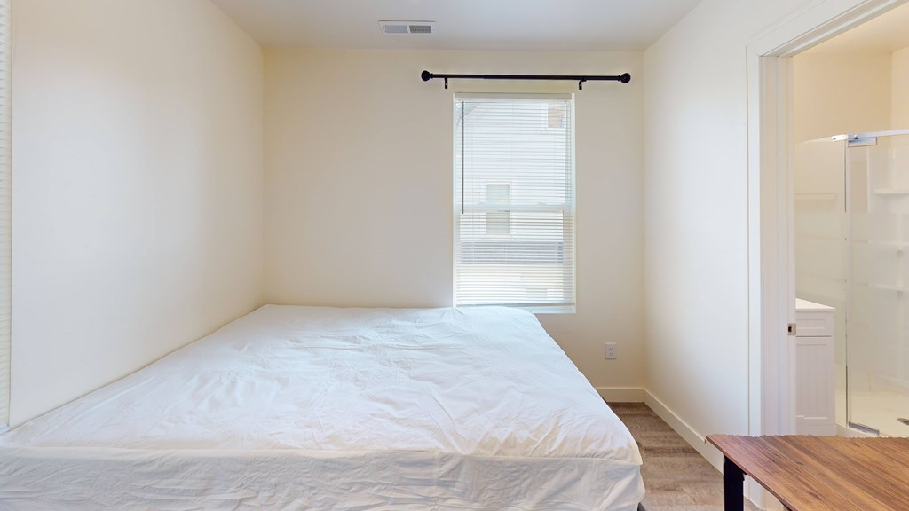 A bedroom with a bed, a window with blinds, a wooden table, and a glass shower stall.